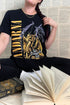 Women sitting crossed legged wearing black Fourth Wing Andarna T-shirt with golden Andarna and purple Tairn dragon and “The Dreamer Sleeps” quote, styled with open book and braided hair.