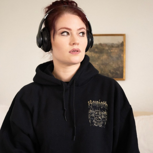 Model in headphones wearing the black Basgiath War College hoodie, small gold War College crest on the front chest, Fourth Wing merch inspired by Rebecca Yarros