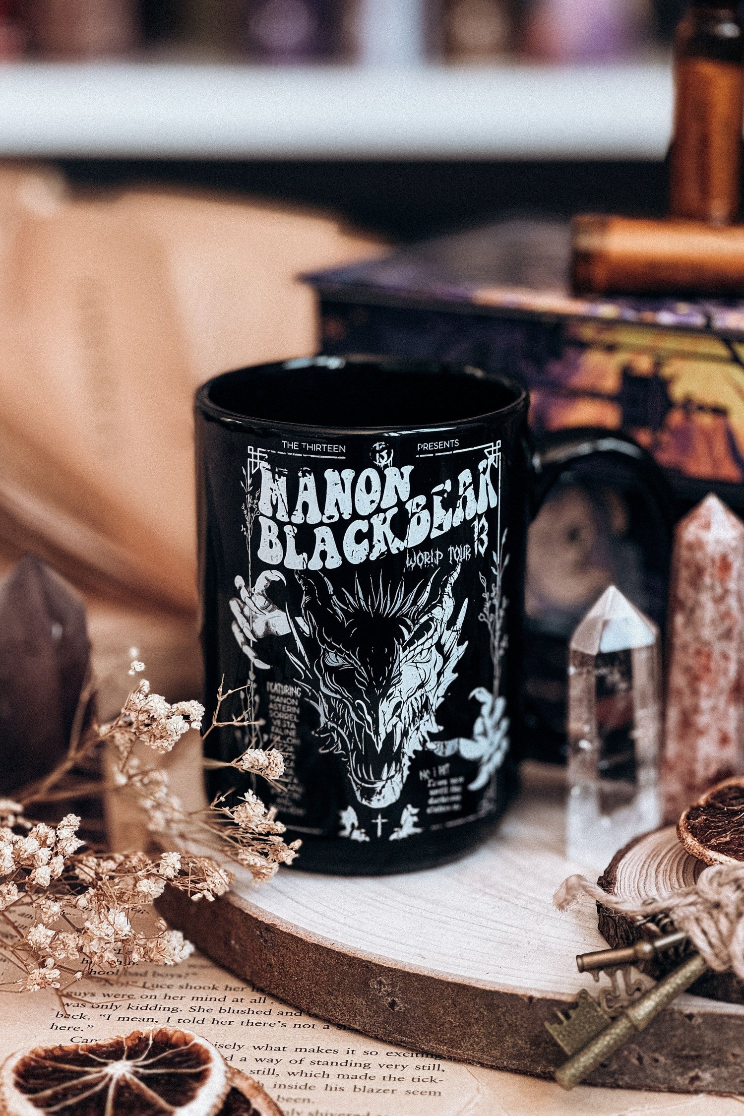 Black ceramic mug with a fierce illustration of Manon Blackbeak, styled with crystals, dried flowers, and a dark fantasy aesthetic on a wooden base.