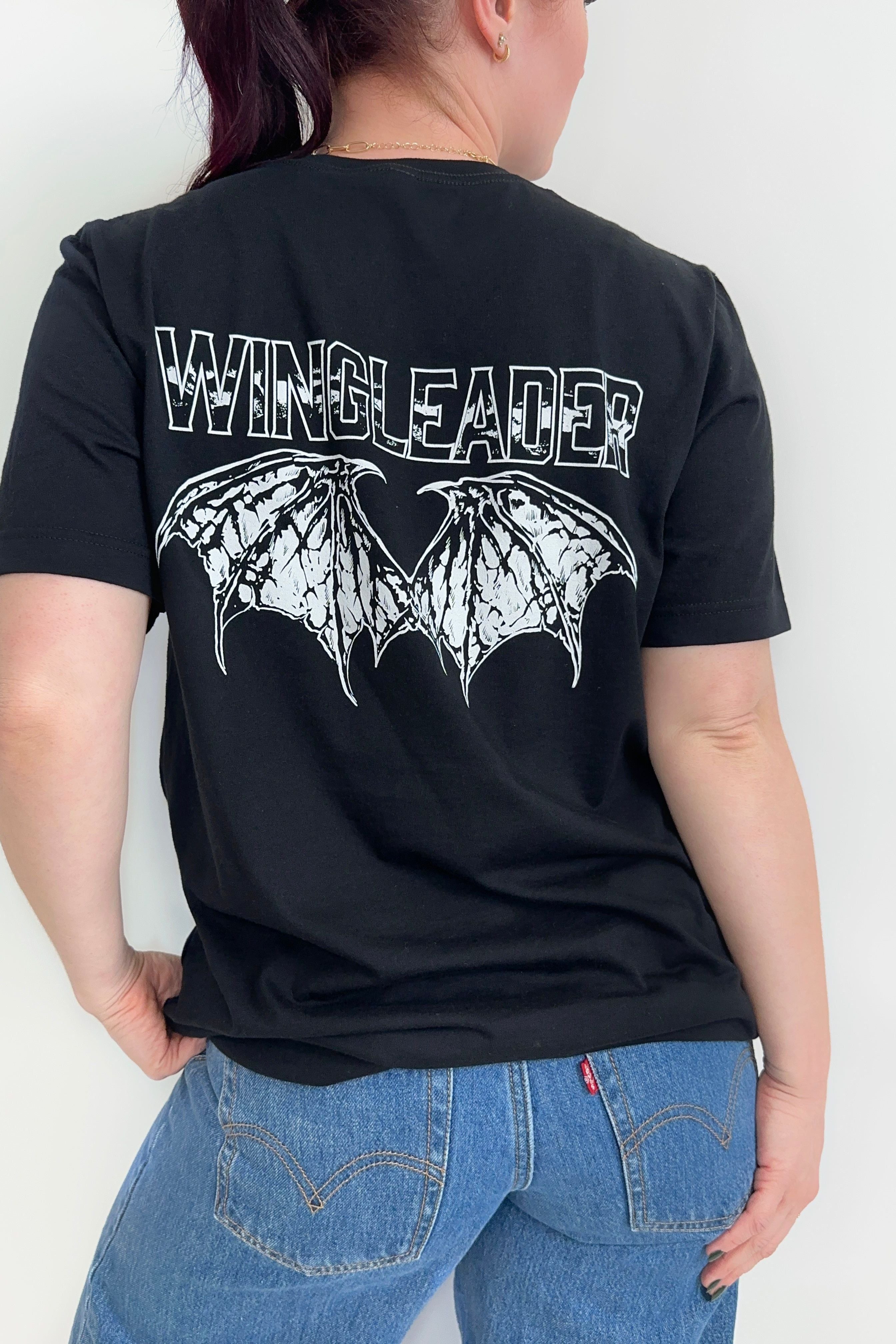 Back view of a black unisex t-shirt with “Wingleader” text and white wingspan illustration in distressed print, worn with jeans