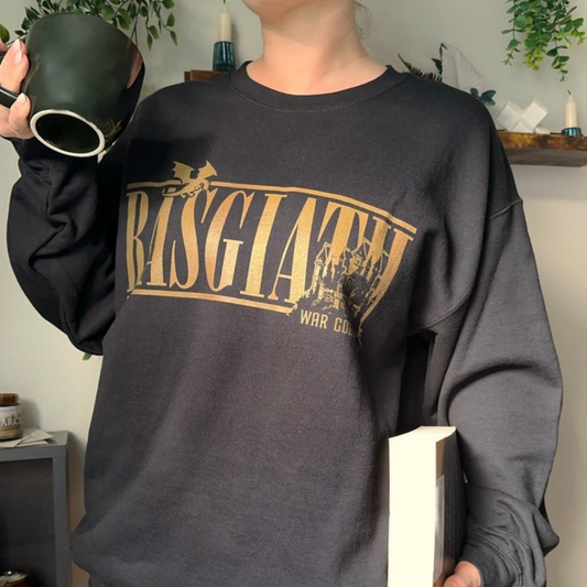Black Basgiath Location sweatshirt held with a mug and book, gold Basgiath lettering with a dragon and War College castle, Fourth Wing merch inspired by Rebecca Yarros