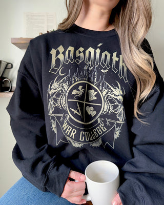 Woman wearing black Basgiath War College Fourth Wing sweatshirt, holding a white mug while seated casually wearing blue jeans.