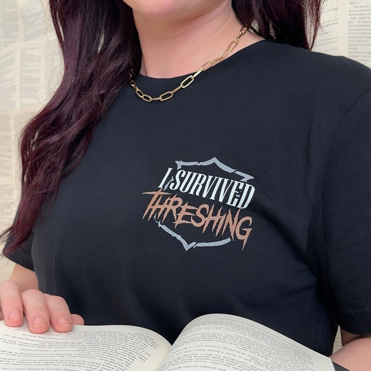 I Survived Threshing t-shirt on a reader holding an open book, black tee with the I Survived Threshing crest on the chest, Fourth Wing