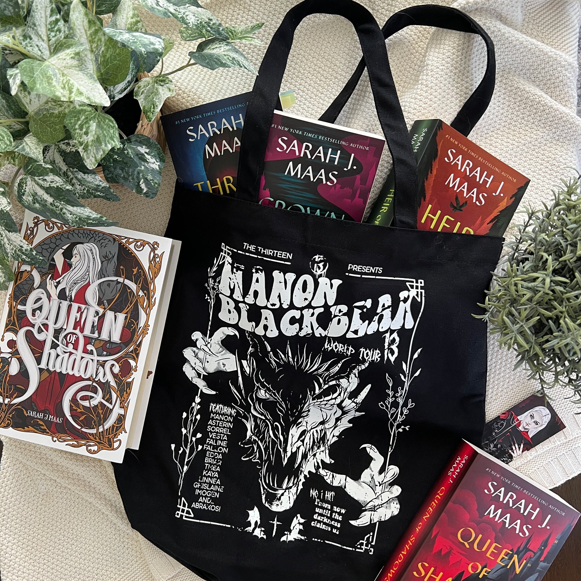Flat-lay of a black Manon Blackbeak tote bag with a white wyvern gig-poster print, styled with Sarah J. Maas hardbacks, inspired by Throne of Glass