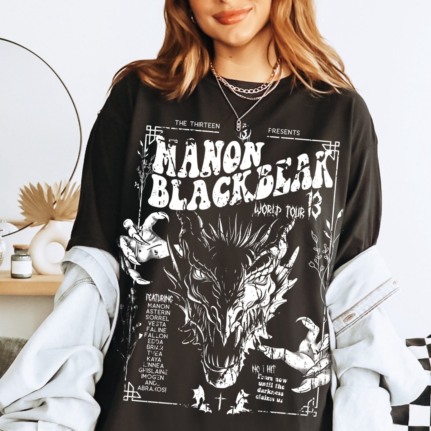 Model in a washed black Manon Blackbeak t-shirt under a pale denim jacket, printed with a white wyvern tour poster, inspired by Throne of Glass by Sarah J. Maas