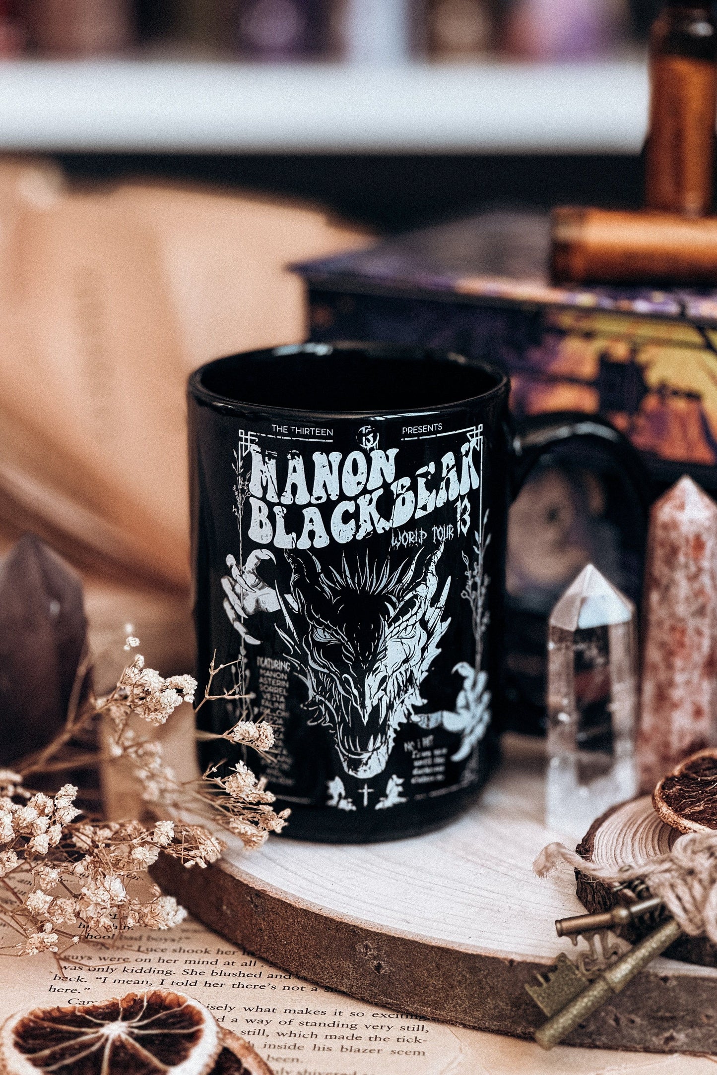 Black ceramic mug with a fierce illustration of Manon Blackbeak, styled with crystals, dried flowers, and a dark fantasy aesthetic on a wooden base.