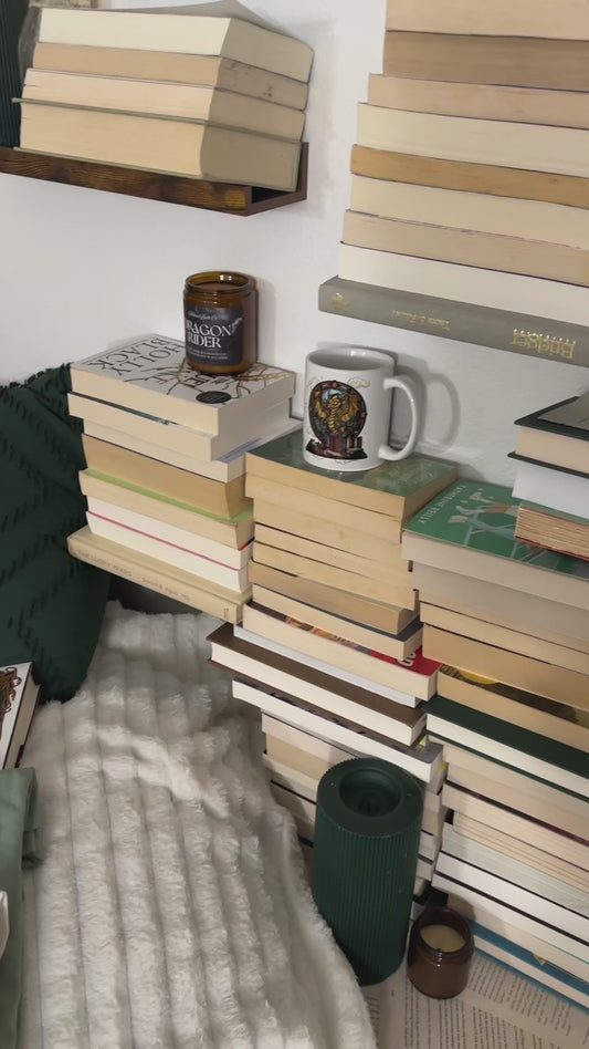 Styled flat lay: the sage and off-white Fireheart tees folded on a bed with Throne of Glass hardbacks, a slow pan across the back print — 'to whatever end', FIREHEART, Aelin Galathynius, 'the world will be saved and remade by the dreamers' [focal 50 60]