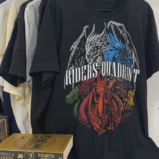 Product video: the black Riders Quadrant t-shirt front print on the rail beside the Fourth Wing hardbacks — the white, blue, red and green dragons in a ring behind the arched RIDERS QUADRANT lettering [focal 50 40]