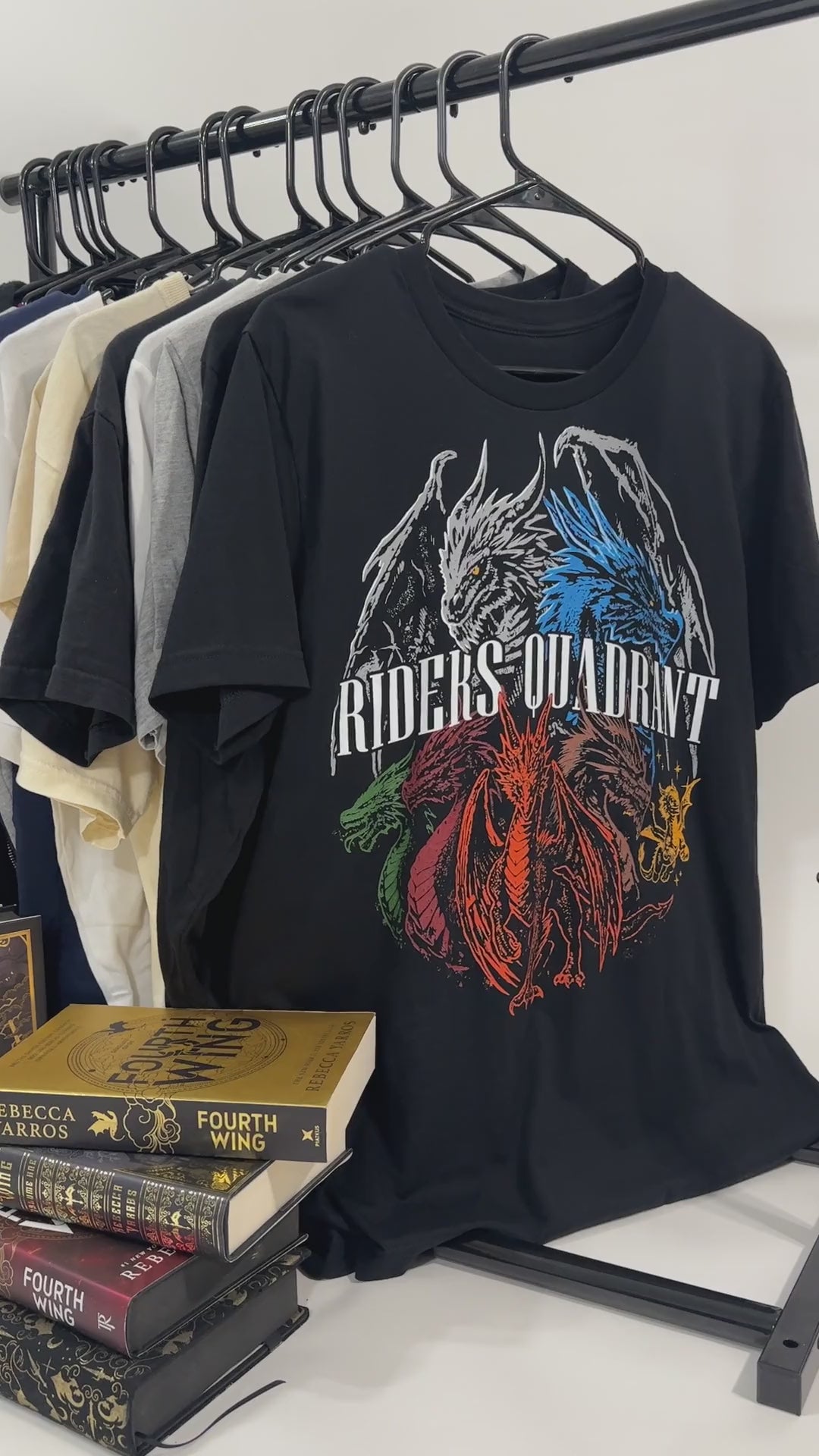 Product video: the black Riders Quadrant t-shirt front print on the rail beside the Fourth Wing hardbacks — the white, blue, red and green dragons in a ring behind the arched RIDERS QUADRANT lettering [focal 50 40]