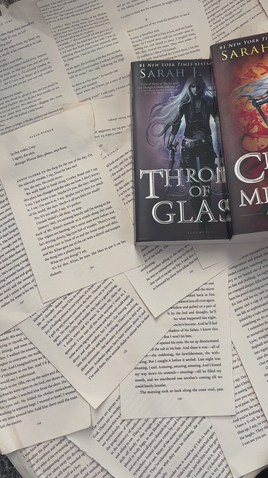Flat lay video: the black Manon Blackbeak t-shirt laid over scattered book pages with the Throne of Glass, Crown of Midnight and Kingdom of Ash hardbacks — the white tour-poster print reading 'The Thirteen presents / Manon Blackbeak / World Tour 13' around the wyvern [focal 50 50]