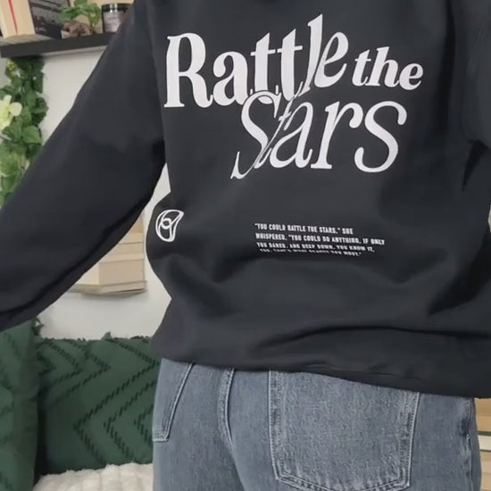 Try-on video: the black Rattle the Stars hoodie — the back print with 'Rattle the' above 'Stars' in a serif lockup, the circular mark and the quote block beneath, then turning to the small chest print [focal 50 20]
