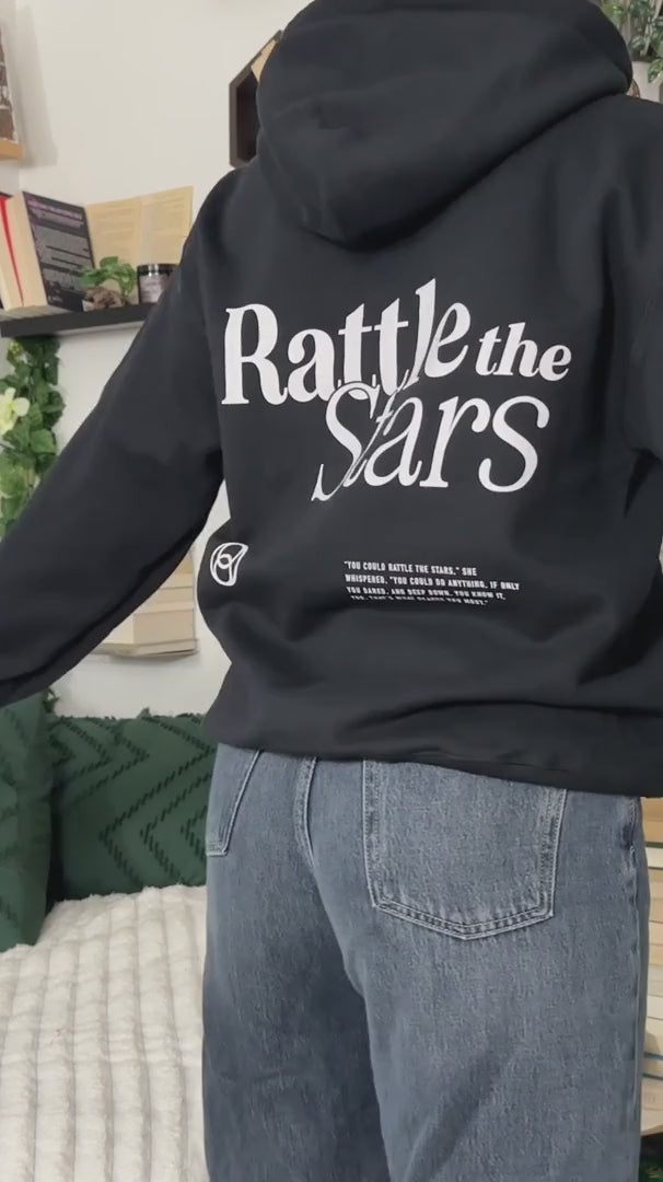Try-on video: the black Rattle the Stars hoodie — the back print with 'Rattle the' above 'Stars' in a serif lockup, the circular mark and the quote block beneath, then turning to the small chest print [focal 50 20]