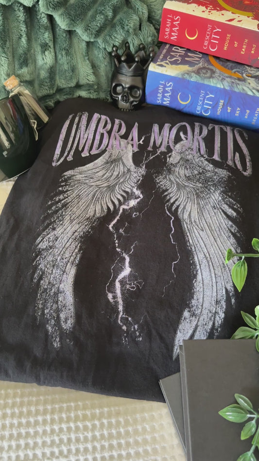 Flat lay video: the black Umbra Mortis t-shirt with the Crescent City books — the arched UMBRA MORTIS lettering above a pair of outstretched wings split by lightning [focal 50 30]