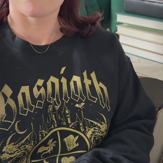 Lifestyle video: the black Basgiath War College sweatshirt worn while reading — the gold gothic Basgiath lettering above the War College crest with its two dragons and banner [focal 50 65]
