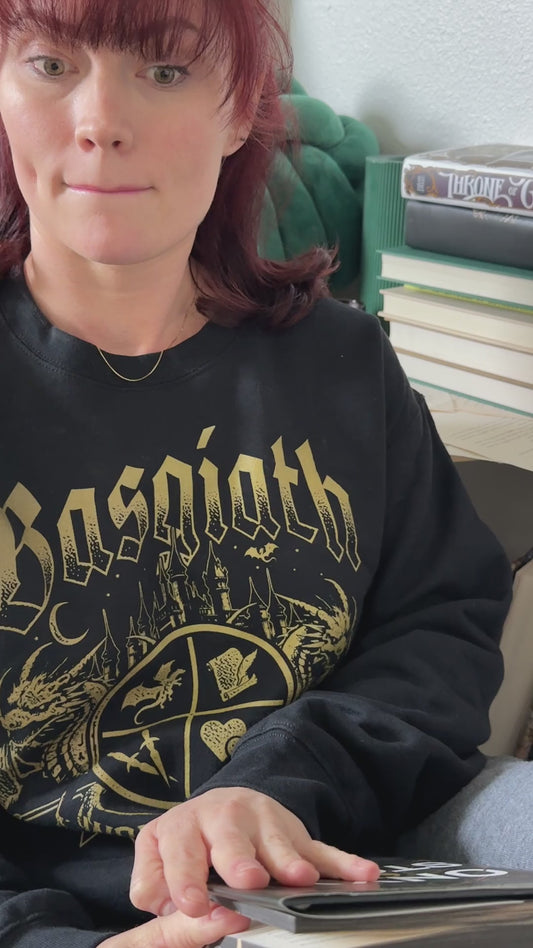 Lifestyle video: the black Basgiath War College sweatshirt worn while reading — the gold gothic Basgiath lettering above the War College crest with its two dragons and banner [focal 50 65]