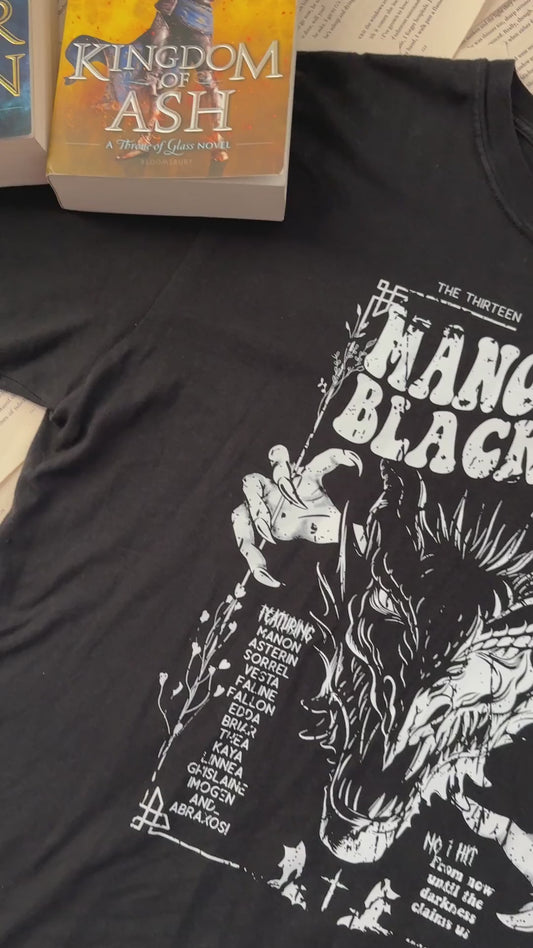 Flat lay video: the black Manon Blackbeak t-shirt with the Throne of Glass hardbacks — the white line-art wyvern under 'THE THIRTEEN PRESENTS / MANON BLACKBEAK / WORLD TOUR 13' in the concert-poster layout [focal 50 40]
