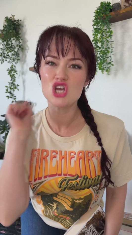 Try-on video: cream Fireheart Festival t-shirt with the retro Beltane poster print, worn with jeans and held beside a Throne of Glass hardback by Sarah J. Maas [focal 50 58]