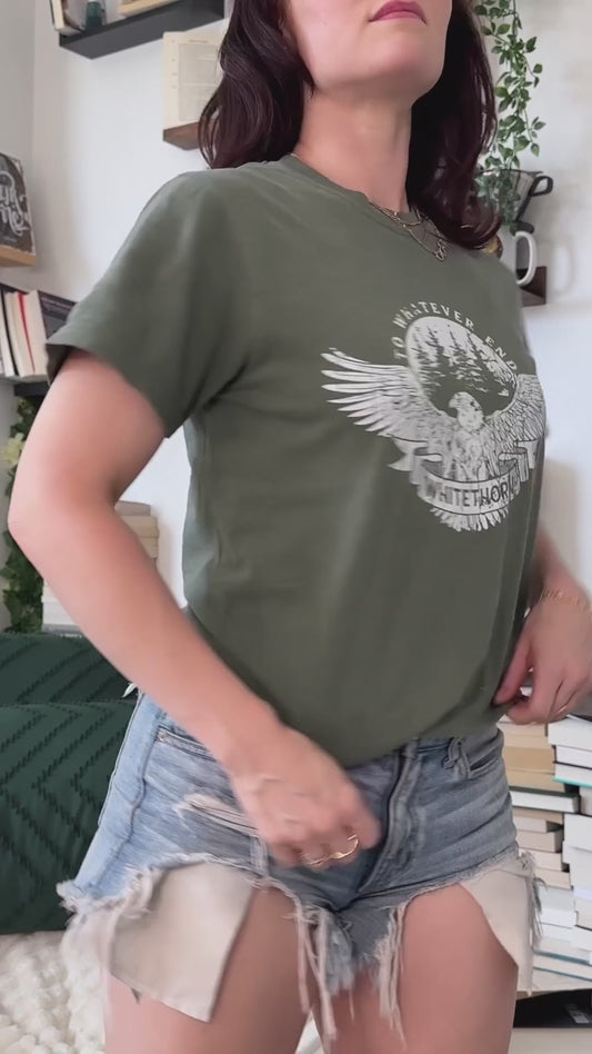 Try-on video: the olive Rowan Whitethorn t-shirt — the circular badge print with 'to whatever end' arched over pines, the eagle with spread wings and WHITETHORN on a banner — then held with a folded stack of the other Throne of Glass tees [focal 50 30]