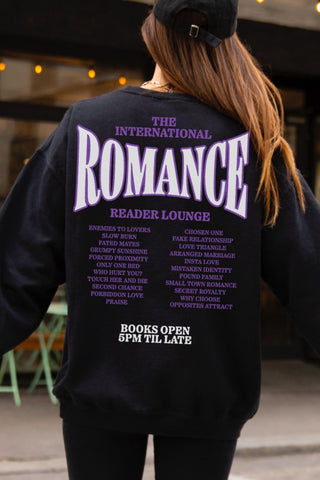 Back view of a women weaing a oversized black sweatshirt with bold lavender text reading “The International Romance Reader Lounge” and a list of romance book tropes like enemies to lovers, grumpy sunshine, and only one bed - designed as cozy book merch for trope-obsessed readers