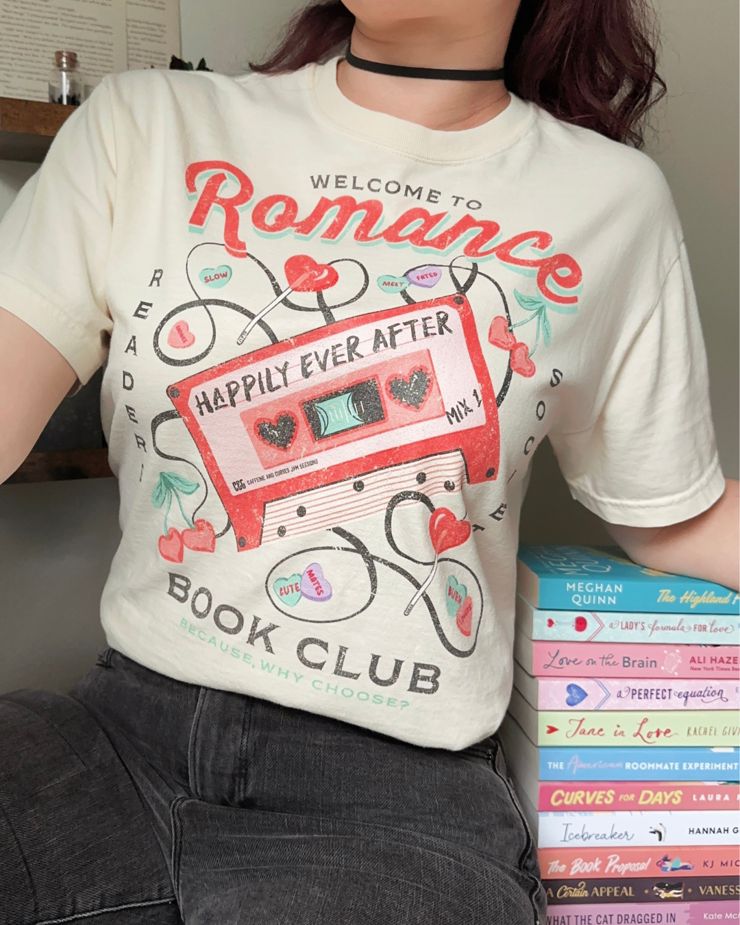 A close-up of a female wearing a cream-colored “Romance Reader Society” t-shirt featuring a retro pink cassette tape labeled ‘Happily Ever After: Mix 1,’ surrounded by candy hearts, headphones, and romance-themed phrases like ‘meet cute,’ ‘slow burn,’ and ‘because… why choose?’ The perfect nostalgic tee for fans of love stories and HEAs.