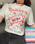 A close-up of a female wearing a cream-colored “Romance Reader Society” t-shirt featuring a retro pink cassette tape labeled ‘Happily Ever After: Mix 1,’ surrounded by candy hearts, headphones, and romance-themed phrases like ‘meet cute,’ ‘slow burn,’ and ‘because… why choose?’ The perfect nostalgic tee for fans of love stories and HEAs.