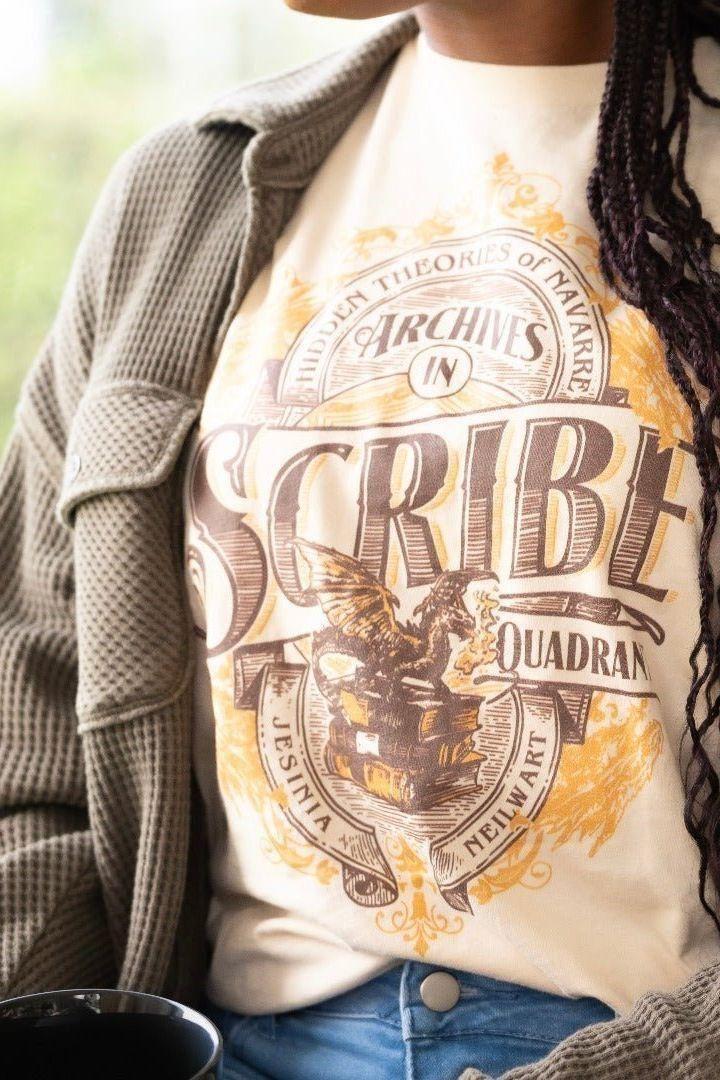 Woman wearing cream Scribe Quadrant Fourth Wing tshirt featuring vintage archive crest, styled with green cardigan, coffee mug and blue jeans.