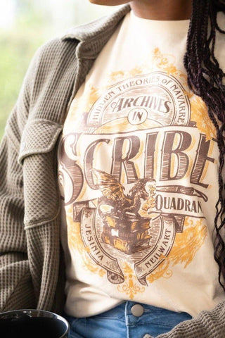 Woman wearing cream Scribe Quadrant Fourth Wing tshirt featuring vintage archive crest, styled with green cardigan, coffee mug and blue jeans.