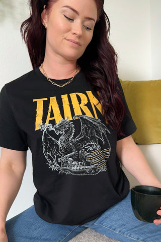 Woman wearing black Fourth Wing Tairn dragon T-shirt, bold yellow lettering spelling 'Tairn', seated casually with coffee cup wearing blue jeans.