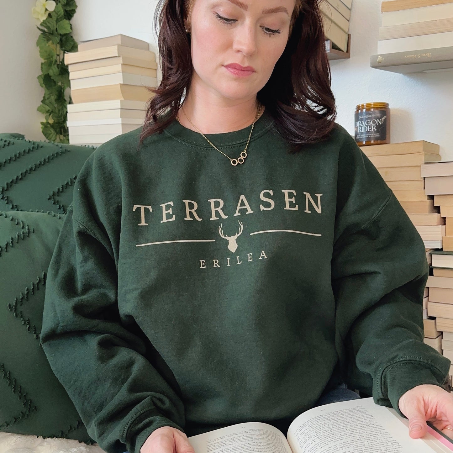 Model reading a book in a forest green Terrasen crewneck printed with TERRASEN, a stag emblem and ERILEA, inspired by Throne of Glass by Sarah J. Maas