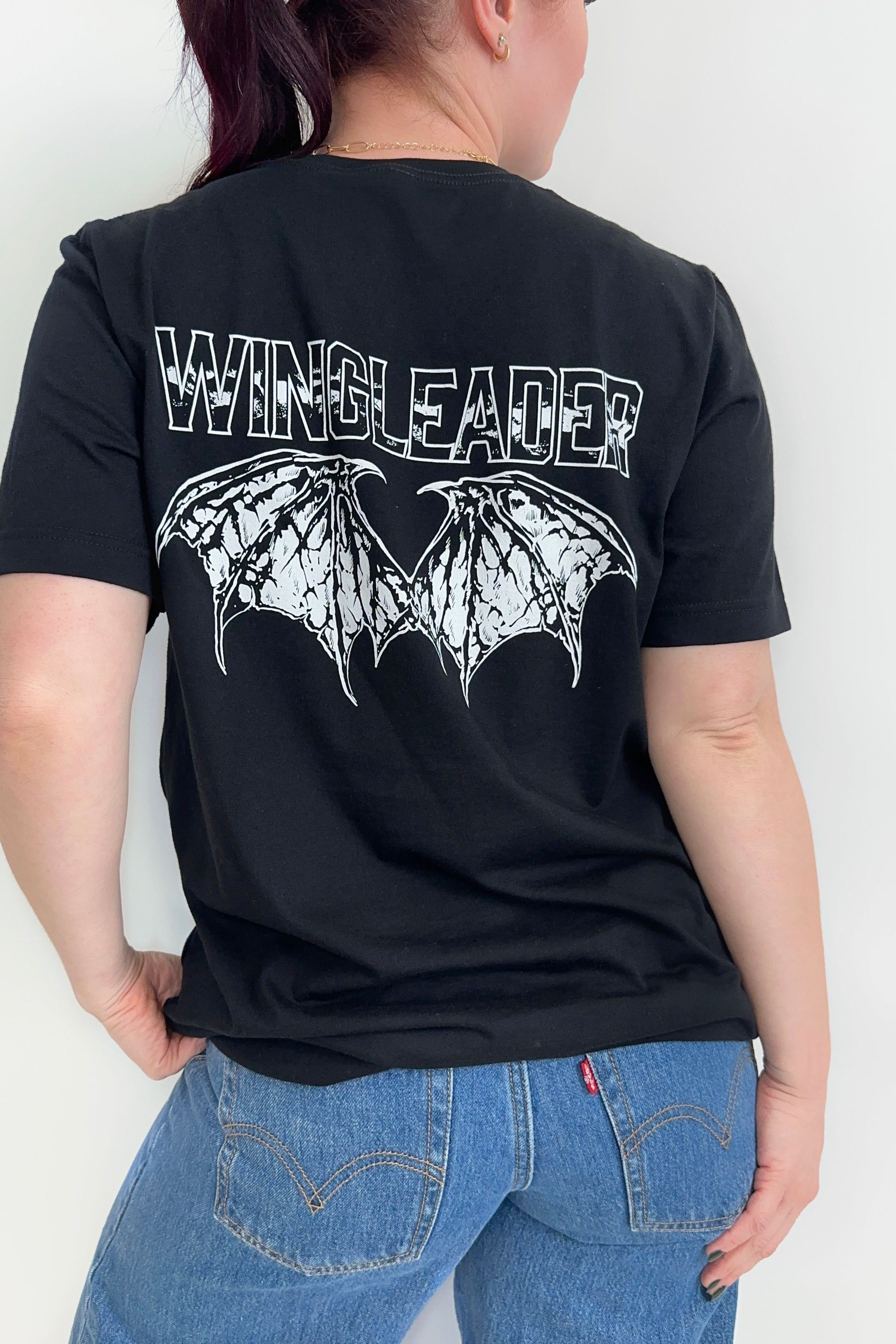 Back view of a black unisex t-shirt with “Wingleader” text and white wingspan illustration in distressed print, worn with jeans